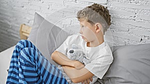 Adorable blond boy in pyjamas, sitting on bed in his bedroom, hugging teddy bear with sad expression, showing signs of unhappiness