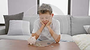 Adorable blond boy, sitting solemn and sad at home, sceptically studying coins and piggy bank, doubting the concept of saving