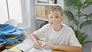 Adorable blond boy, student at his classroom desk, beaming with happiness while drawing in his notebook, surrounded by books