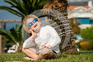 Adorable blond boy in sunglasses and hat on scenic ocean shore sitting on the green gruss