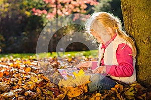 Adorable blond child reading book by the trunk in autumnal sunset afmosphere