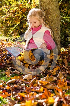 Adorable blond child reading book by the trunk in autumnal sunset afmosphere