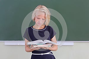 Adorable blond kid girl reading a book in a classroom near a chalkboard. Back to school.