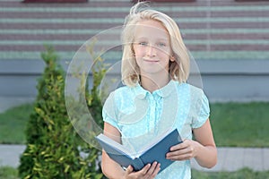 Adorable blonde girl with book in hands near school. Schoolgirl likes to learn and read.