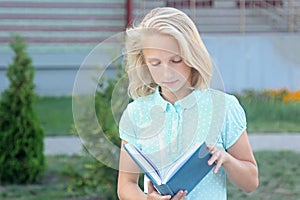 Adorable blonde girl with book in hands near school. Schoolgirl likes to learn and read.