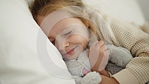 Adorable blonde girl hugging rabbit doll lying on bed at bedroom