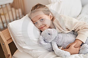 Adorable blonde girl hugging rabbit doll lying on bed at bedroom