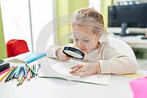 Adorable blonde girl student reading book using magnifying glass at classroom