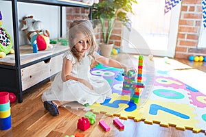 Adorable blonde toddler playing with building blocks toy around lots of toys at kindergarten