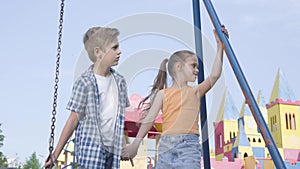 Adorable boy and beautiful girl with long hair holding hands and talking near the swing, smiling happily. A couple of