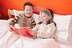 Adorable boy and girl reading book sitting on bed at bedroom