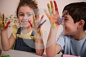 Adorable boy and girl showing their colorfully painted hands in gouaches while enjoying art class