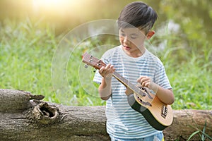 Adorable boy with guitar sitting on the grass on sunset, Musical concept with little boy playing ukulele at sunny park