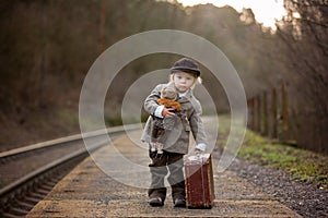 Adorable boy on a railway station, waiting for the train with suitcase and beautiful vintage doll