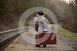 Adorable boy on a railway station, waiting for the train with suitcase and beautiful vintage doll