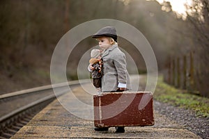 Adorable boy on a railway station, waiting for the train with suitcase and beautiful vintage doll