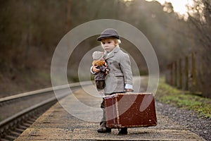 Adorable boy on a railway station, waiting for the train with suitcase and beautiful vintage doll