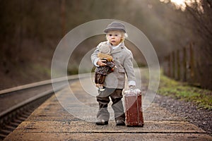 Adorable boy on a railway station, waiting for the train with suitcase and beautiful vintage doll