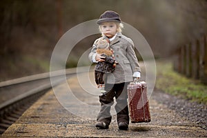 Adorable boy on a railway station, waiting for the train with suitcase and beautiful vintage doll