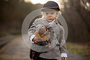 Adorable boy on a railway station, waiting for the train with suitcase and beautiful vintage doll