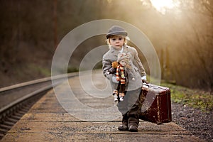 Adorable boy on a railway station, waiting for the train with suitcase and beautiful vintage doll