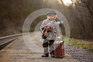 Adorable boy on a railway station, waiting for the train with suitcase and beautiful vintage doll