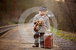 Adorable boy on a railway station, waiting for the train with suitcase and beautiful vintage doll