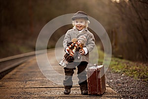 Adorable boy on a railway station, waiting for the train with suitcase and beautiful vintage doll