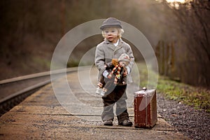 Adorable boy on a railway station, waiting for the train with suitcase and beautiful vintage doll