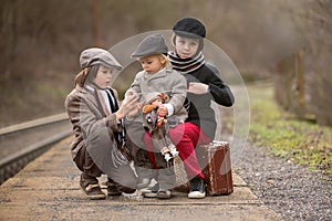 Adorable boy on a railway station, waiting for the train with suitcase and beautiful vintage doll