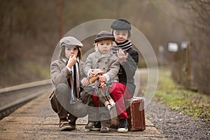 Adorable boy on a railway station, waiting for the train with suitcase and beautiful vintage doll