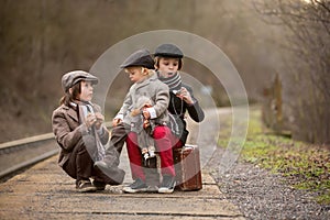 Adorable boy on a railway station, waiting for the train with suitcase and beautiful vintage doll