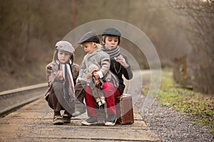 Adorable boy on a railway station, waiting for the train with suitcase and beautiful vintage doll