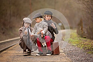 Adorable boy on a railway station, waiting for the train with suitcase and beautiful vintage doll