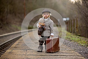 Adorable boy on a railway station, waiting for the train with suitcase and beautiful vintage doll