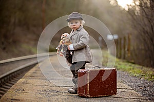 Adorable boy on a railway station, waiting for the train with suitcase and beautiful vintage doll