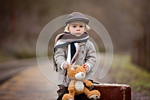 Adorable boy on a railway station, waiting for the train with suitcase and sweet teddy bear