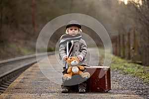 Adorable boy on a railway station, waiting for the train with suitcase and sweet teddy bear