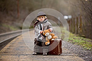 Adorable boy on a railway station, waiting for the train with suitcase and sweet teddy bear