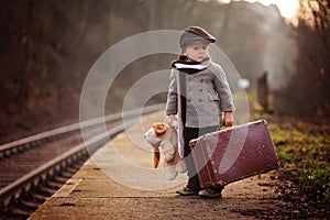 Adorable boy on a railway station, waiting for the train with suitcase and teddy bear