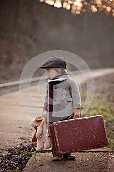 Adorable boy on a railway station, waiting for the train with suitcase and teddy bear