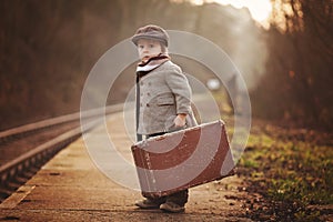 Adorable boy on a railway station, waiting for the train with suitcase and teddy bear