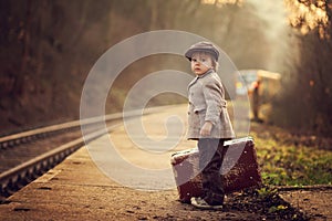 Adorable boy on a railway station, waiting for the train with suitcase and teddy bear