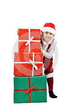 Adorable boy in santa clothes peeks out behind Christmas big gift boxs. Isolated white background.