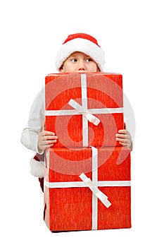 Adorable boy in santa clothes peeks out behind Christmas big gift boxs. Isolated white background.