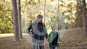 Adorable boy is walking in forest with his loving father holding his hand, bearded young man is talking and gesturing