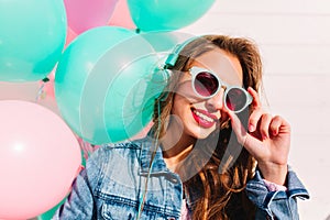 Adorable brunette young woman looking through stylish sunglasses and posing with smile next to colorful balloons. Close