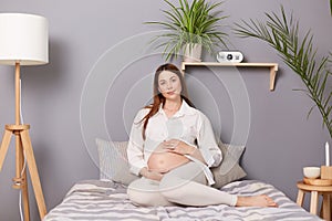 Adorable calm pregnant brown haired caucasian woman sitting in bed resting embracing her belly looking at camera resting at home