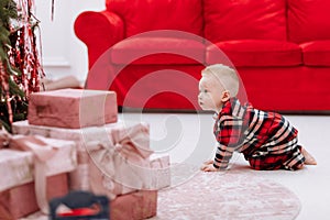 Adorable caucasian baby boy is crawling by the Christmas tree, many festive gift boxed and red sofa at home, cute infant