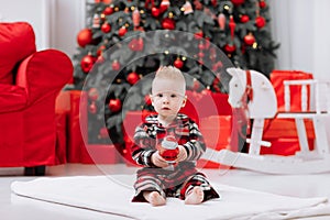 Adorable caucasian baby boy is sitting and holding gift toy Santa Claus near a Christmas tree, many festive gift boxed